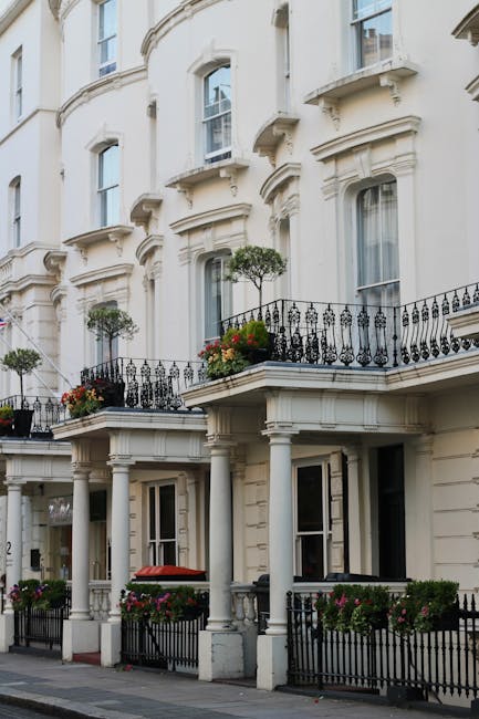 A close-up view of a mature tree with thick, textured branches and lush green leaves in the foreground, set against a red brick residential building featuring classical sash windows and small black me